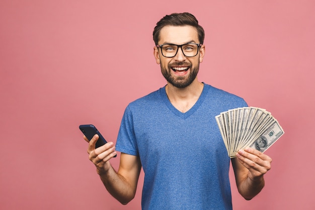 Excited man in casual t-shirt holding lots of money in dollar currencys and cell phone in hands isolated over pink wall.