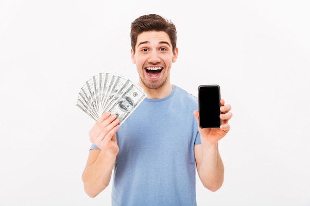 Excited man in casual t-shirt holding lots of money in dollar currencys and cell phone in hands, isolated over white wall