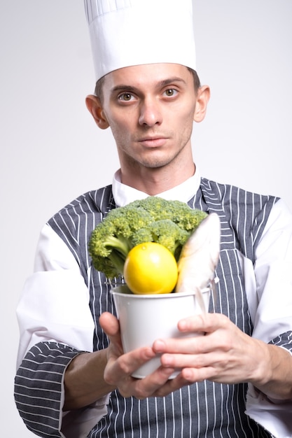 制服を着て興奮した男のシェフ料理人は、白い背景の上に分離された野菜のバケツを保持します