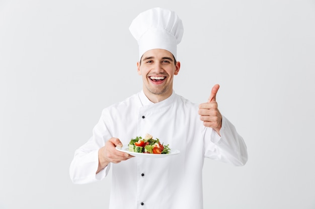 Excited man chef cook wearing uniform showing fresh green salad on a plate isolated over white wall, thumbs up