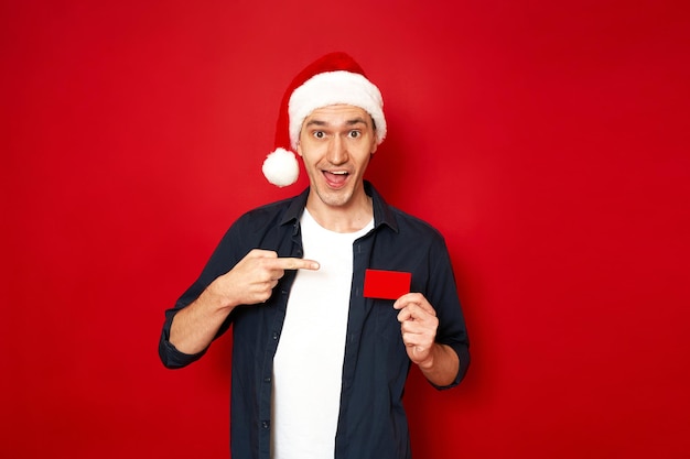 Excited man in Christmas hat shows credit plastic bank card isolated red background space for text