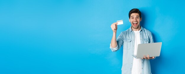 Excited man shop online showing credit card and holding laptop buying in internet standing over person