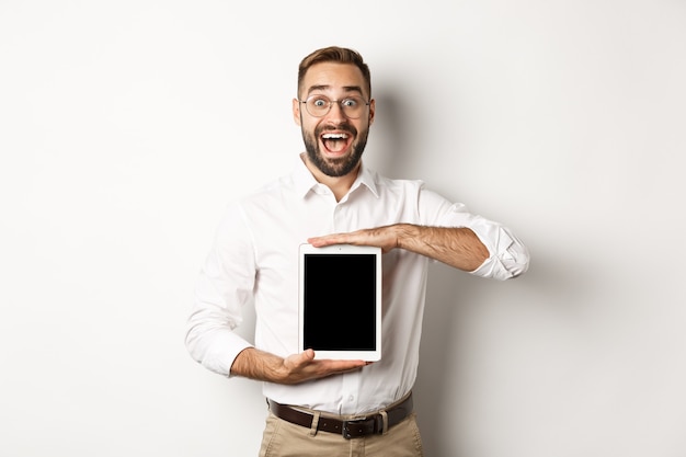 Взволнованный человек показывает экран цифрового планшета, изумленно улыбаясь, стоя на белом фоне.