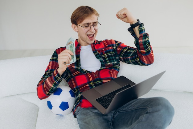 집에 앉아 축구 경기 축하 승리를 보고 있는 흥분된 남자 현금 돈과 축구 공을 들고 있는 남자 감정적인 남자가 온라인 스포츠 베팅에서 이깁니다