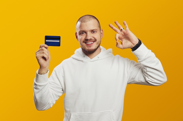 Excited man in white hoodie holding credit card while showing ok gesture isolated over yellow wall