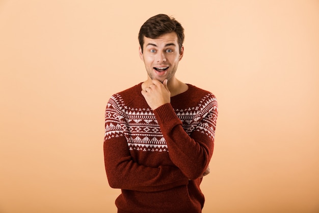 excited man with bristle wearing knitted sweater posing on camera and touching chin, isolated over beige wall