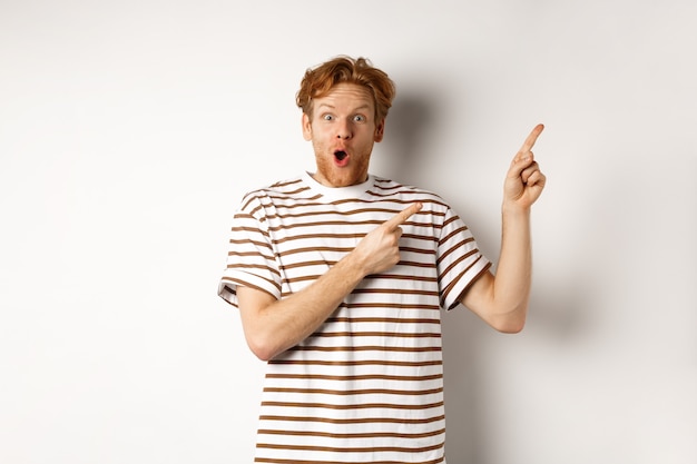 Excited man with curly red hair showing promo offer and looking amazed, pointing fingers at upper right corner logo, white background.