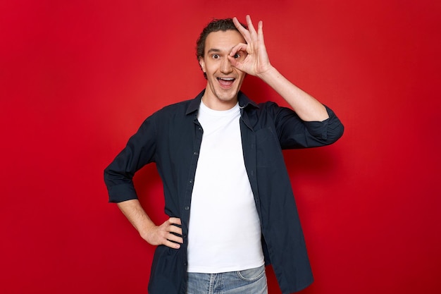 Excited man with mouth open holds his hand in form of an ok sign near eye and looks through fingers