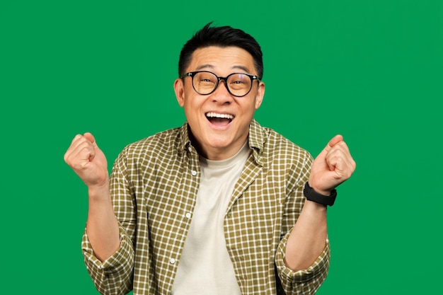 Photo excited mature asian man screaming in excitement and gesturing yes with both hands posing on green