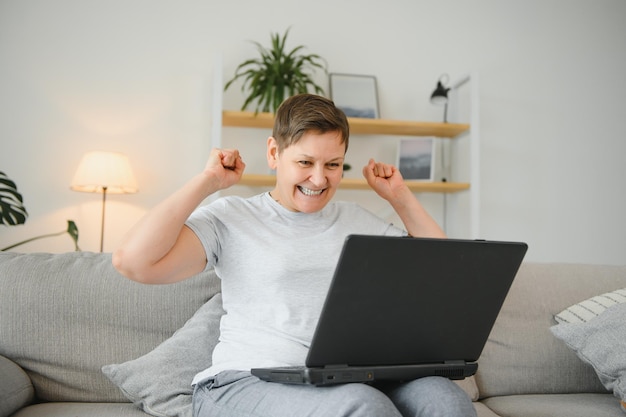 Excited mature woman looking at laptop screen reading good news in message celebrating online lottery win rejoicing success overjoyed older female sitting on couch at home using computer