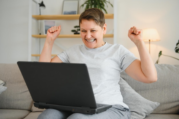 Excited mature woman looking at laptop screen reading good news in message celebrating online lottery win rejoicing success overjoyed older female sitting on couch at home using computer