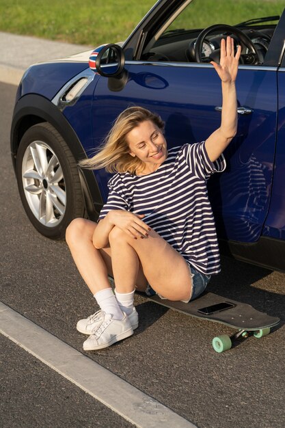 Eccitato donna di mezza età agitando la mano saluto amico seduto su longboard fuori dall'auto in campagna