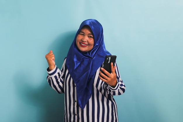 Excited middleaged Asian woman in hijab is holding a mobile phone isolated on a blue background