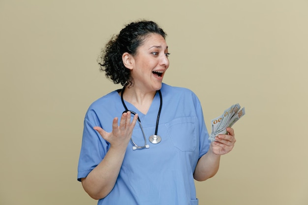 excited middleaged female doctor wearing uniform and stethoscope around neck holding money looking at it showing empty hand isolated on olive background