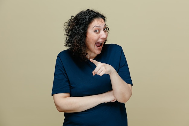 Excited middleaged woman wearing tshirt looking at camera pointing to side isolated on olive green background