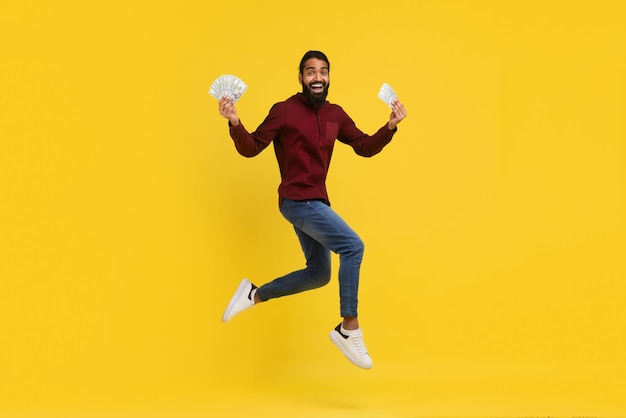 Excited millennial indian guy holding money cash in hands jumping