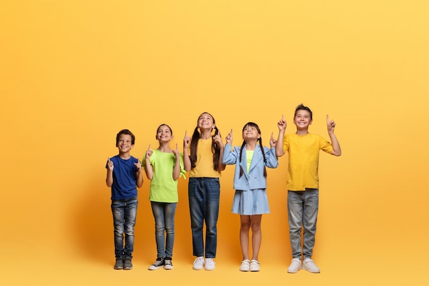 Excited multiracial children pointing up at blank space yellow background