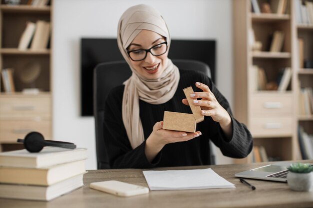 Excited muslim woman sitting at desk and opening parcel box