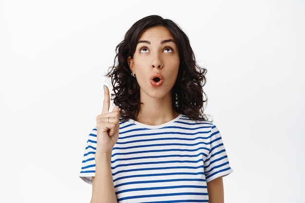 Excited natural girl pointing, looking up with impressed face, say wow, checking out special promo deal, showing advertisement logo on top, standing against white background.