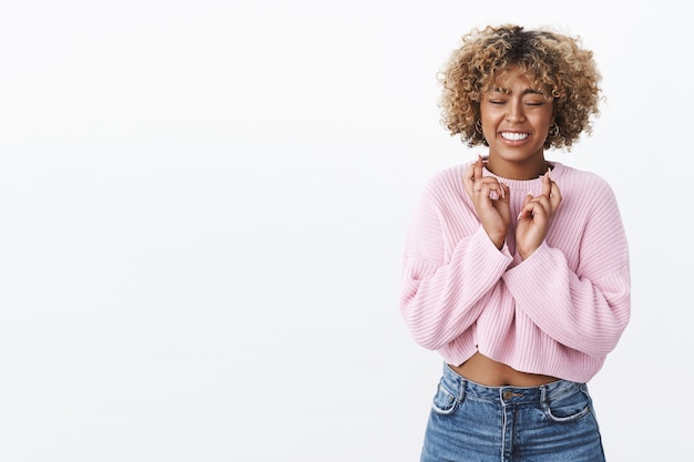 Eccitata e nervosa donna afro-americana carina che esprime desiderio e sorride incrociando le dita per buona fortuna spera che il sogno diventi realtà posa entusiasta con gli occhi chiusi e un sorriso speranzoso contro il muro bianco