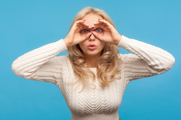 Excited nosy blond woman in white sweater looking through holes in fingers like binoculars, spying, trying to find out secrets, private detective. Indoor studio shot isolated on blue background