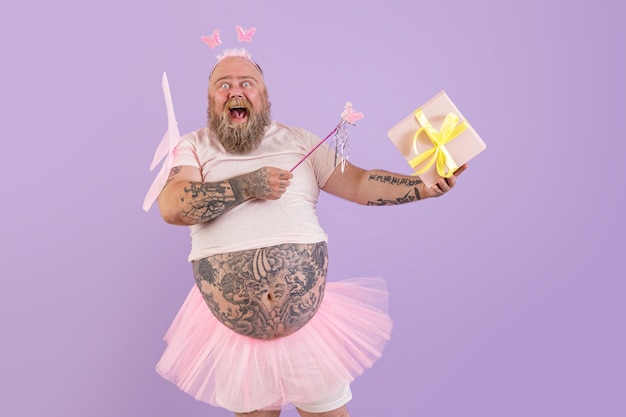 Excited obese male person in fairy costume holds gift box on\
purple background