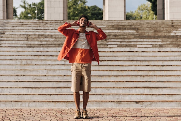 Excited optimistic brunet man in beige shorts and orange jacket smiles sincerely listens to music in red headphones outside