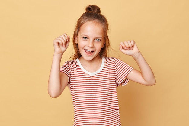 Excited overjoyed little girl wearing striped tshirt standing isolated over beige background screaming with happy facial expression clenched fists