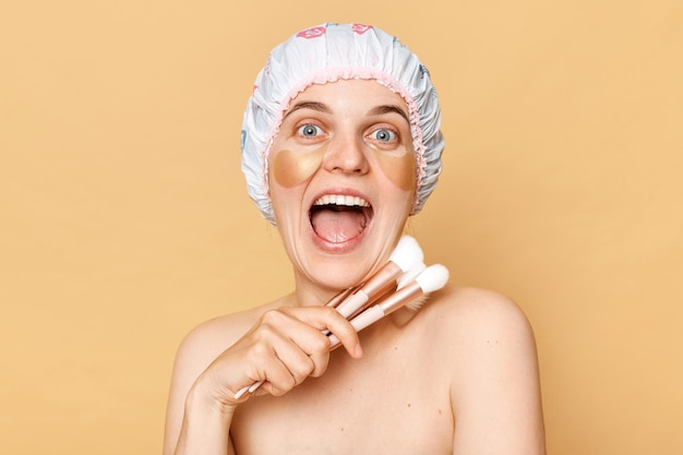 Excited overjoyed woman holding makeup face brushes and use shower cap to keep hair looking at camera with amazed look standing isolated over beige background