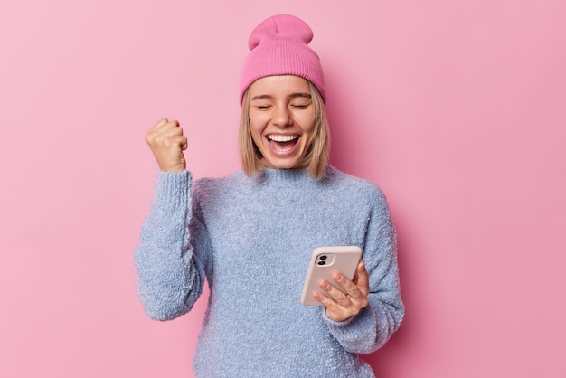 Excited overjoyed woman holds mobile phone and does winner gesture celebrates winning lottery triumphs over success dressed in casual jumper and hat isolated over pink background Yes I did it