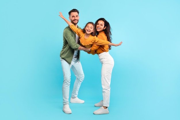 Excited parents playing with daughter holding her in arms posing on blue background girl spreading