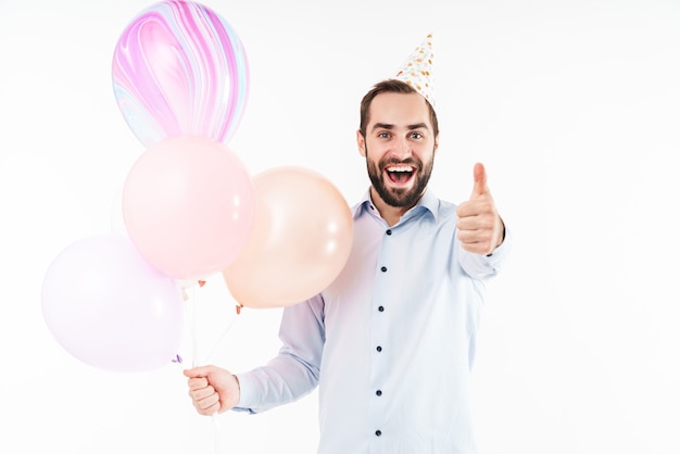 excited party man holding air balloons and gesturing thumb up isolated over white wall