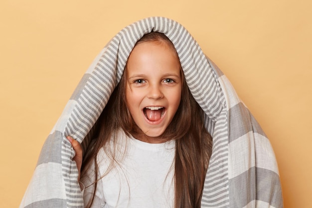 Excited positive little girl with blanket standing isolated over beige background screaming with happy facial expression yelling loud frighting