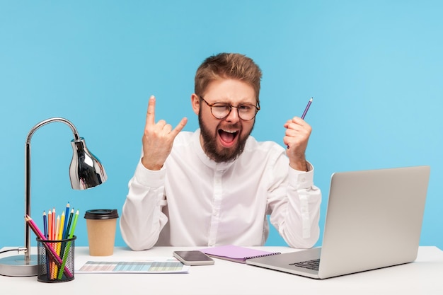 Excited positive man office worker showing rock and roll gesture sitting at workplace with laptop notepad and pencil successfully finished annual report. Indoor studio shot isolated on blue background