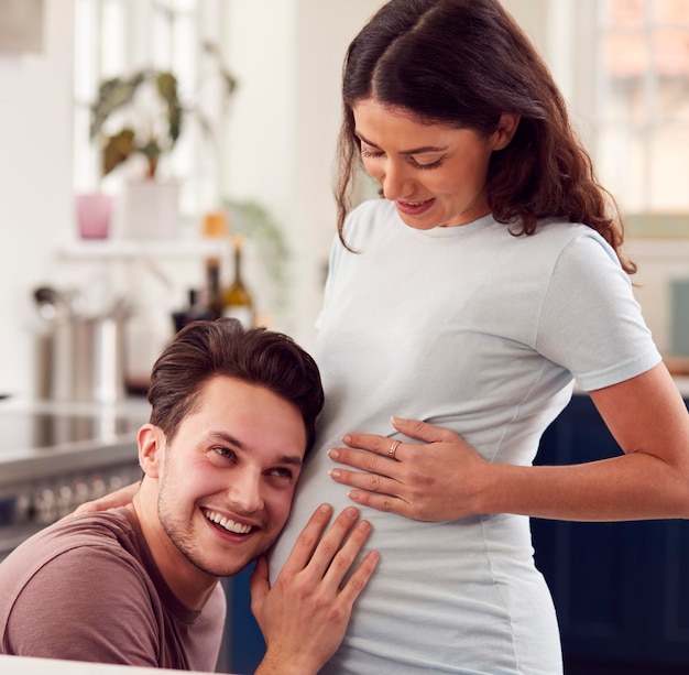 Excited Pregnant Transgender Couple At Home In Kitchen With Man Listening To Baby39s Heartbeat