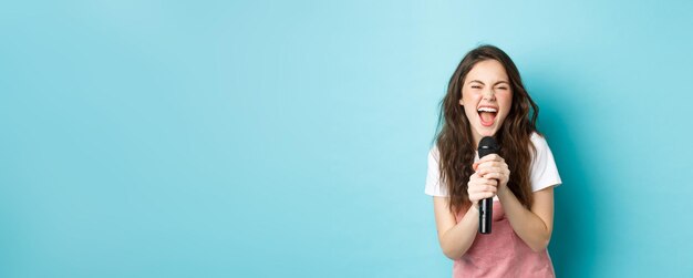 Excited pretty girl singing karaoke holding microphone and smiling happy standing over blue background person