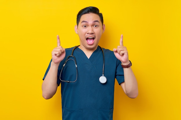 Excited professional young Asian male doctor or nurse wearing a blue uniform and stethoscope pointing finger up on copy space isolated on yellow background Healthcare medicine concept