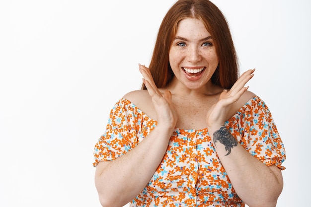 Excited redhead girl claps hands and smiles looking amazed at camera standing over white background