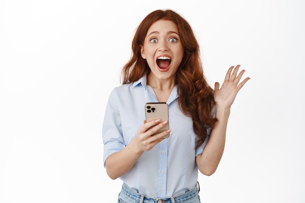 Excited redhead girl screams from amazement and surprised, holding smartphone, react to awesome discounts, great news online, celebrating winning money on phone, white background