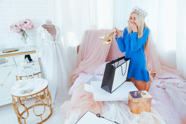 Photo excited rich girl after shopping in flamboyant bedroom. blonde lady in tiara on bed unpacking dresses and heeled shoes.
