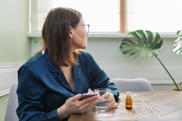 Donna matura triste emozionante che legge smartphone. donna in pigiama seduta a casa, acqua potabile con gocce sedative. salute mentale fisica e psicologica delle persone di mezza età, copia spazio