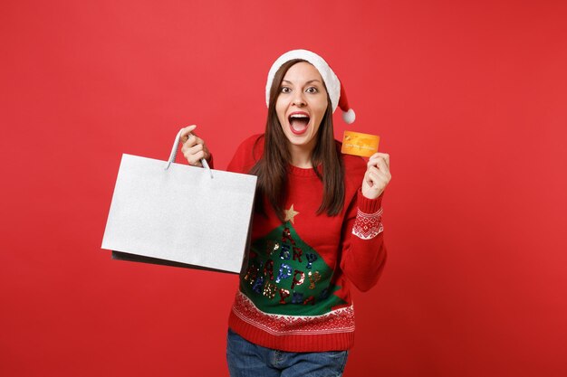 Photo excited santa girl keeping mouth wide open, holding credit card, packages bags with purchases after shopping isolated on bright red background. happy new year 2019 celebration holiday party concept.