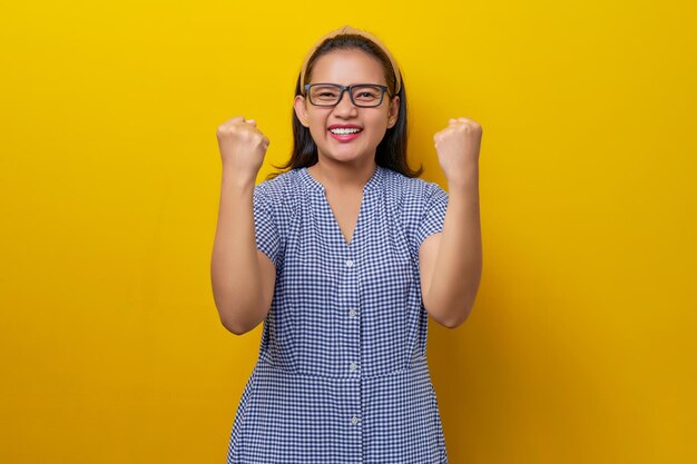 Excited satisfied young Asian woman wearing a dress checkered with glasses raising her fists up celebrating success isolated on yellow background people lifestyle concept