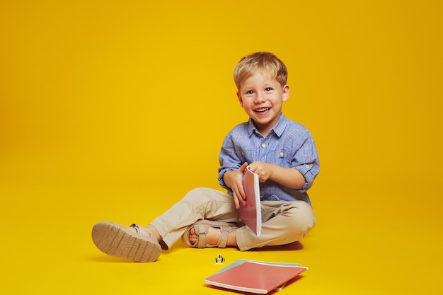 Photo excited schoolboy sitting on floor and holding notebook while happily smiling at camera