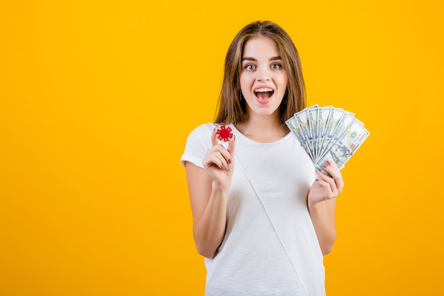 Excited screaming brunette girl with red poker chip and one hundred dollar bills in hand isolated over yellow