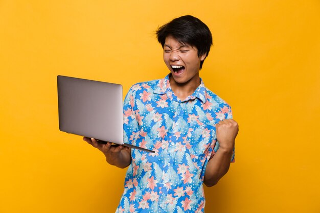 Excited screaming young asian man standing isolated over yellow space using laptop computer make winner gesture.