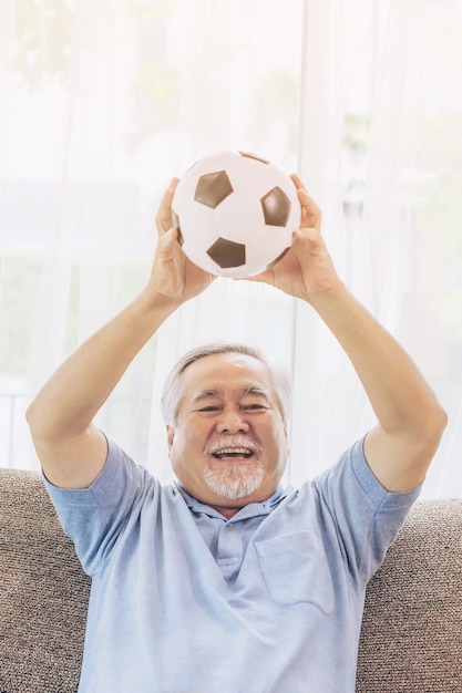 Eccitato uomo anziano, il vecchio si sente felice tenendo in mano il pallone da calcio prepararsi per il preferito della squadra di cheer sul divano del divano a casa, lo stile di vita senior asiatico concetto di buona salute