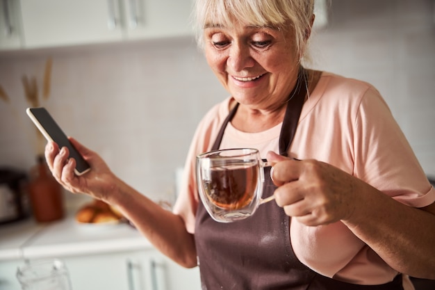 Donna anziana eccitata che mangia tè caldo nella sua cucina
