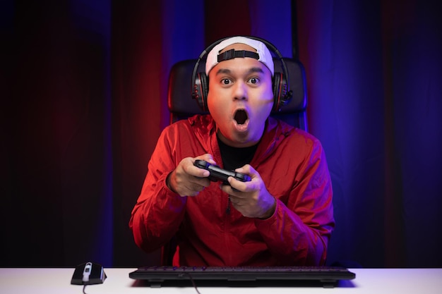 Excited and shocked face of asian gamer with headphone holding\
joystick playing video game online sitting on chair at living room.\
indian professional gamer streaming on social playing game very\
fun