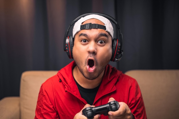 Excited and shocked face of Asian gamer with headphone holding joystick playing video game online sitting on sofa at living room Indian professional gamer streaming on social playing game very fun